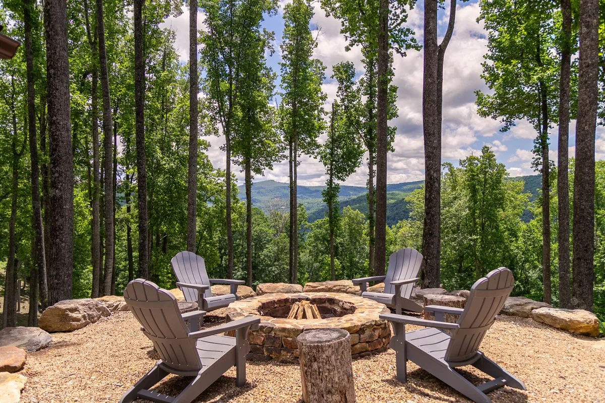 This image captures a tranquil fire pit area with wooden chairs arranged in a semi-circle, surrounded by lush trees and featuring a sweeping mountain view in the background. The serene setting is perfect for gatherings, stargazing, or enjoying a peaceful mountain evening.