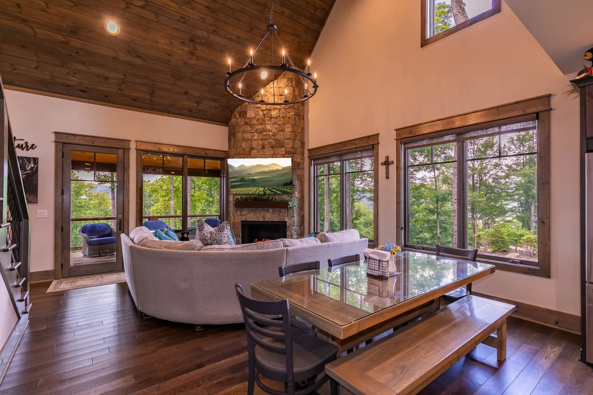 This image showcases a spacious open-plan living area where a large sectional sofa faces a beautiful stone fireplace and a 65" TV. Large windows along the wall allow natural light to pour in, illuminating the space and offering glimpses of the surrounding mountain scenery. The dining table is visible in the background, further emphasizing the connected gathering spaces.