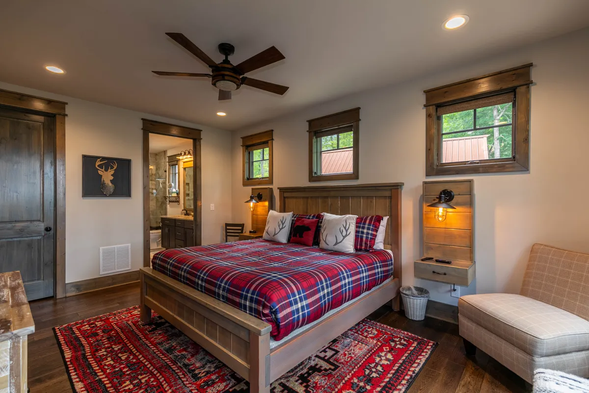 A cozy bedroom featuring a wooden bed frame dressed in a plaid bedspread, a decorative rug, wall art with a deer motif, and a window offering views of surrounding nature. The room exudes a rustic, restful atmosphere, ideal for relaxation after a day of adventure.
