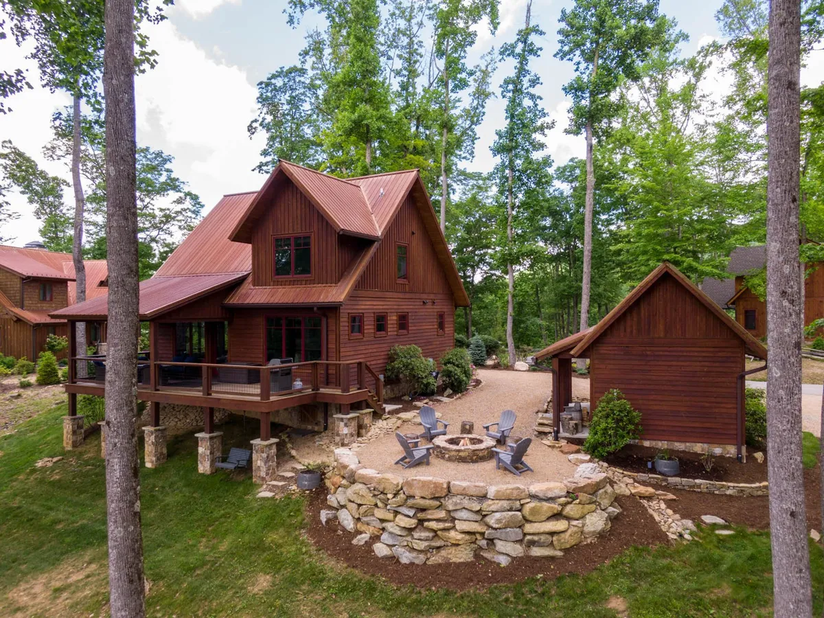 The image showcases a charming wooden cabin nestled among tall trees, featuring a spacious deck and a stone firepit surrounded by comfortable seating. The setting is tranquil, with lush forest all around, inviting guests to gather outdoors, relax, and soak in mountain views.