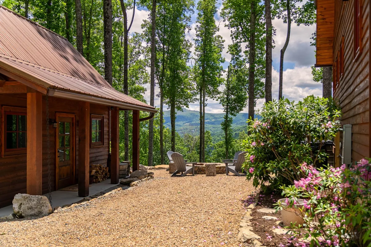 A rustic wooden cabin sits among lush greenery, with Adirondack chairs and a stone-ringed fire pit positioned to take in a sweeping mountain view. The scene is peaceful and inviting, ideal for relaxing, storytelling, and enjoying nature.