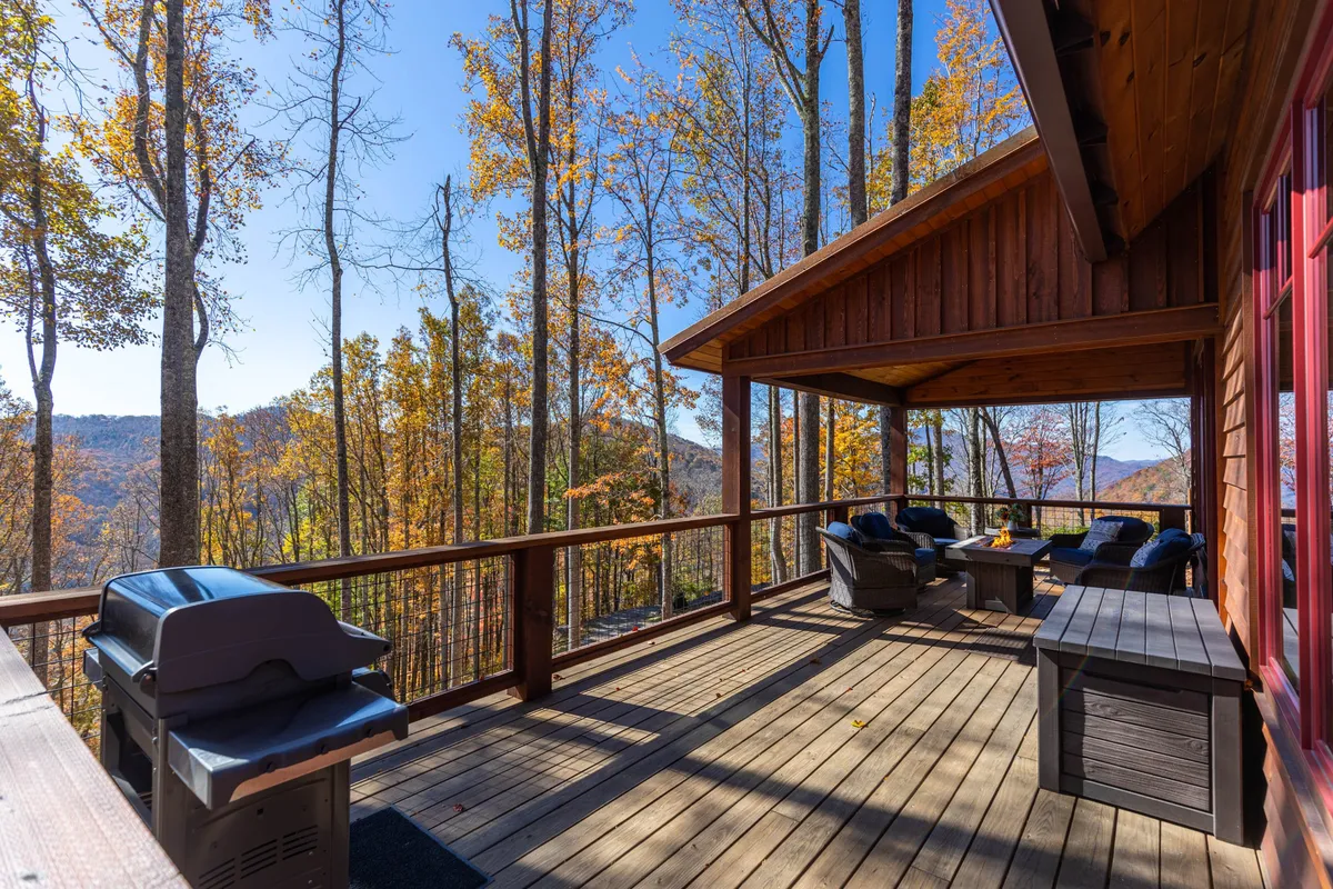 A spacious wooden deck with comfortable seating, a grill for alfresco meals, and sweeping mountain views framed by surrounding trees. The partially covered section provides shelter, while the open area invites guests to enjoy the fresh air and scenery.