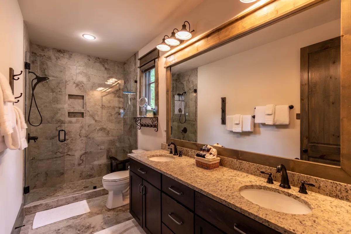 A modern bathroom with a double sink granite vanity, a large mirror, and a spacious walk-in glass-walled shower. The space is bright, clean, and spa-inspired, offering boutique hotel-style comfort.