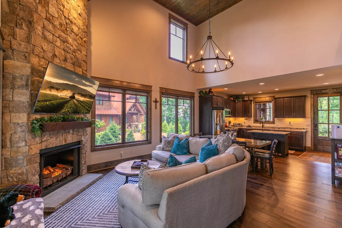This inviting space features a stone fireplace, a comfy sofa with decorative cushions, a dining area, and expansive windows with sunset views. The warm, welcoming atmosphere encourages relaxation, conversation, and togetherness.