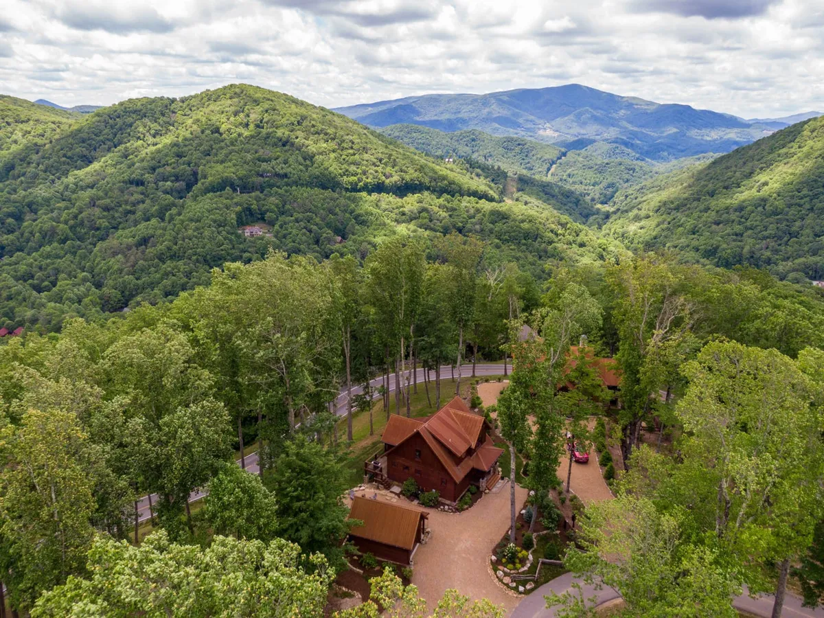 A wide-angle view of a charming wooden cabin nestled among the mountains, with a lush tree canopy and a winding road leading up to the property. The expansive setting highlights the surrounding natural landscape, ideal for outdoor exploration and adventure.