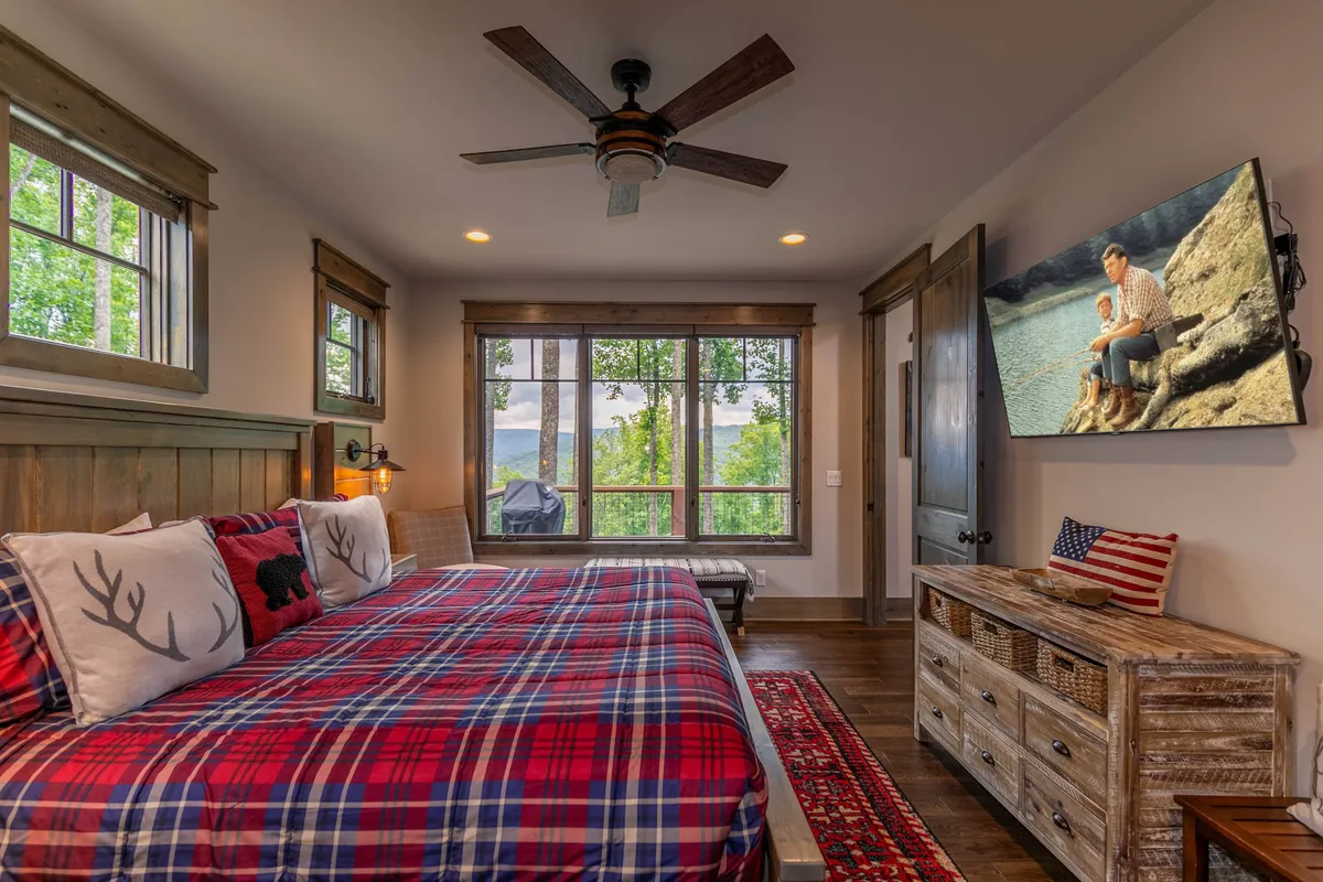 A comfortable bedroom featuring a plaid bedspread, large windows that flood the room with light, a SmartTV mounted on the wall, and a wooden dresser. The mood is relaxed and inviting, perfect for unwinding with a movie or soaking in the morning view.