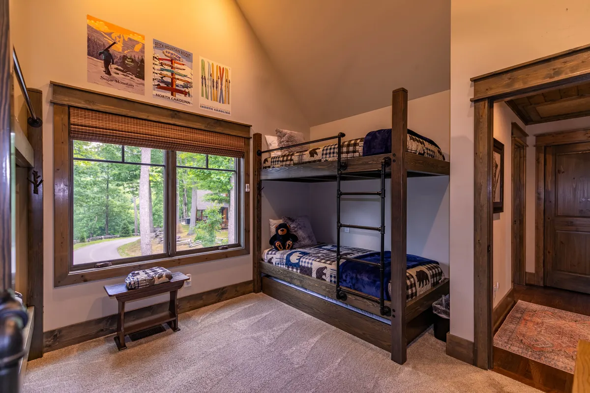 A bright, playful bunk room with sturdy wooden bunk beds, a large window letting in natural light, colorful decorative posters, and practical storage furniture. The space is clearly designed for children or groups, offering both fun and comfort.