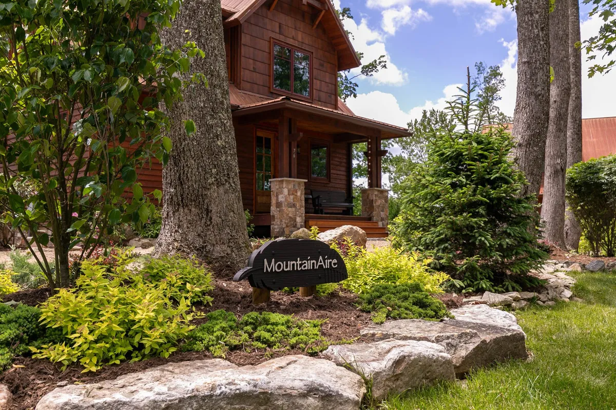 The exterior of MountainAire is featured, including the house facade, the prominent 'MountainAire' sign, and manicured landscaping. The welcoming porch is visible, and the surrounding natural scenery hints at open areas for outdoor activities.
