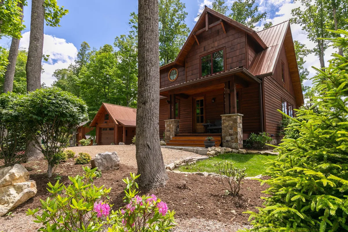 A welcoming view of a wooden house with a spacious front porch, manicured garden, and mature trees all around. The scene captures the sense of a family-friendly mountain retreat, with ample outdoor space for children and pets to play or for gatherings on the porch.