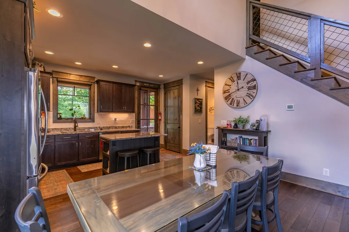 A well-lit view of the kitchen and dining area, showing the kitchen island, a large dining table with seating for eight, and a staircase leading to the upper level. The space is airy, modern, and perfect for shared meals or celebrations.